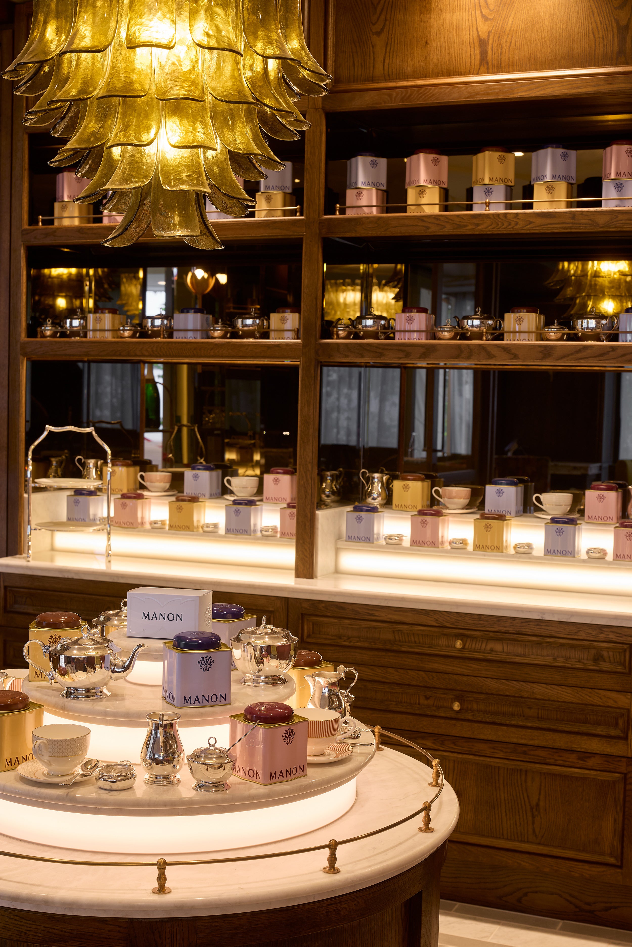 A warm, elegant tea display in a luxury tea room or shop, featuring a round marble table with teapots, cups, and branded MANON tea tins on a glowing white circular base. In the background, wooden shelves are lined with numerous tea boxes and glassware under warm amber lighting.