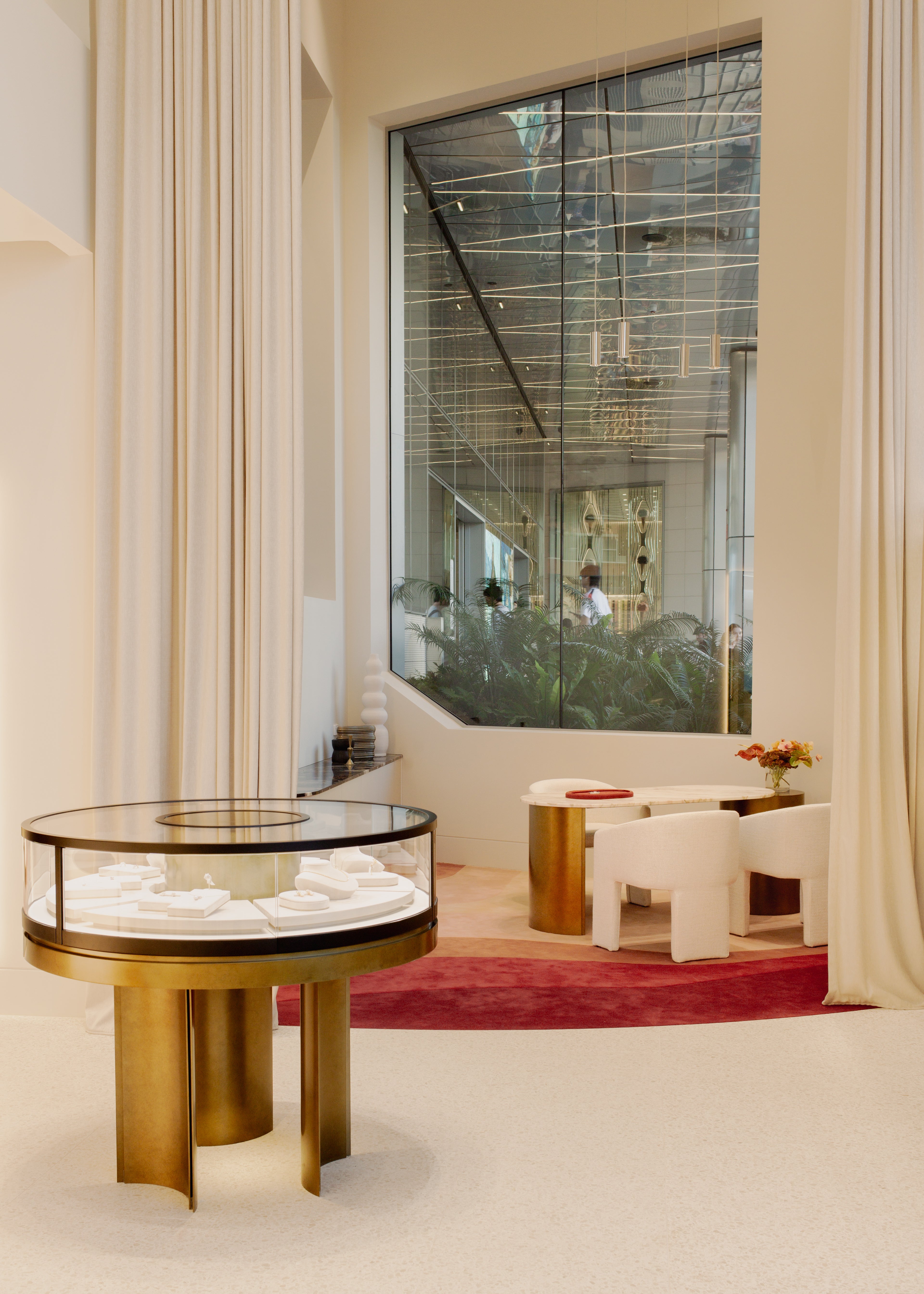 A luxury display case with a round glass top and gold-toned metal legs, showcasing white jewelry, set in a modern, elegant interior with cream walls, draped curtains, and a large window overlooking greenery.